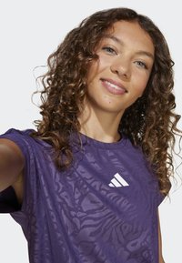 Woman with curly brown hair wearing a purple Adidas sleeveless shirt, smiling and taking a selfie against a plain light background.