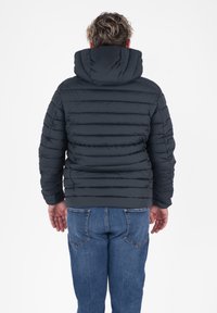 Uomo di spalle che indossa una giacca nera imbottita con cappuccio e jeans blu su uno sfondo bianco.