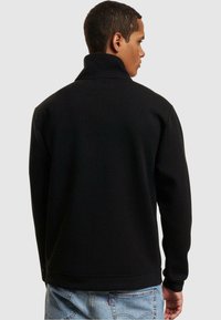 Sort fleece-pullover med høj krave, featuring lange ærmer og en løs pasform, parret med lyseblå denimjeans.