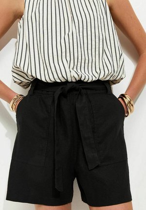 Kobieta nosząca czarne krótkie spodenki z wysokim stanem i przewiązanym paskiem oraz biały bezrękawnik z pionowymi czarnymi paskami, uzupełniona złotymi bransoletkami.