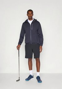 Casaco leve masculino azul-marinho com bolsos com fecho, combinado com calções pretos e ténis atléticos azul-marinho; em pé com um taco de golfe.