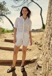 Camicia bianca in pizzo e pantaloni corti con bordi scallop; abbinati a sandali neri con punta aperta e dettagli intrecciati. Ambientazione naturale all'aperto.