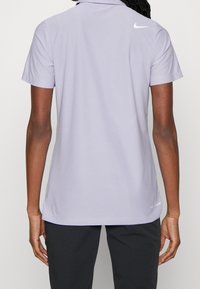 Světle fialové polo tričko Nike s límcem, krátkými rukávy a bočními rozparky. Na zadní straně je bílé logo a tričko je vyrobeno z hladkého, lehkého materiálu.