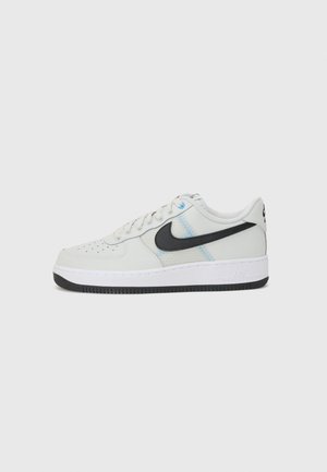 Biały niskoprofilowy sneaker Nike Air Force 1 z czarnym logo Swoosh, perforowanym przodem i czarną podeszwą na jasnoszarym tle.