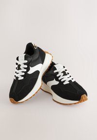 Paire de baskets athlétiques noir et blanc avec des lacets blancs et des semelles en caoutchouc marron texturées sur un fond clair uni.