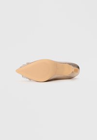Kengät korotettuina, metallinhohtoisella pronssisella yläosalla, terävä kärki ja kukkakristallikoriste kantapäässä. Pohja on teksturoitu ja beige.