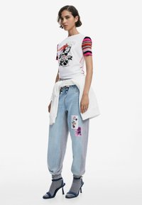 White t-shirt with black and red graphic, striped sleeves in pink and red. Light blue jeans with patch details. Heeled sandals with blue accents.