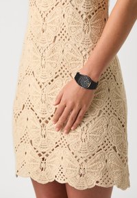 Montre noire avec un cadran rond, bracelet en caoutchouc et accents argentés, associée à une robe en crochet beige avec des motifs de papillons et un ourlet festonné.