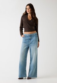 Maglione corto marrone con scollatura a ondine abbinato a pantaloni a gamba larga blu chiaro. I pantaloni hanno un lavaggio sbiadito e tasche laterali.