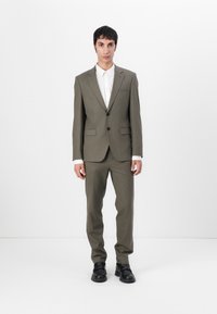Olive green suit with a tailored jacket and straight-leg trousers, paired with a white shirt. Smooth texture, minimalist design, and black footwear.