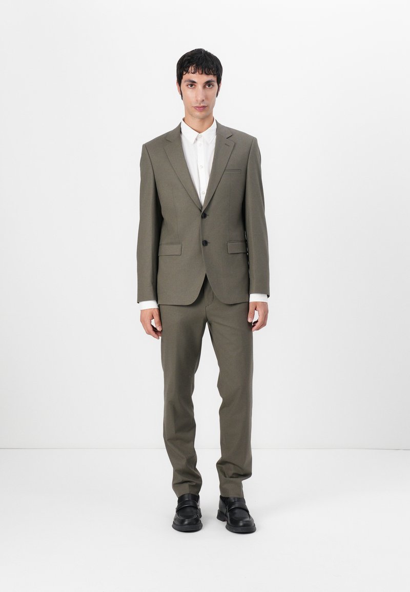 Olive green suit with a tailored jacket and straight-leg trousers, paired with a white shirt. Smooth texture, minimalist design, and black footwear.