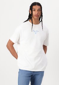 Λευκό βαμβακερό μπλουζάκι με χαλαρή εφαρμογή, στρογγυλή λαιμόκοψη και ανοιχτό μπλε τυπωμένο κείμενο στο στήθος. Συνδυάζεται με μπλε τζιν παντελόνι.