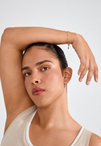 Kuldne käevõru, millel on kettlinkide disain, kantud randmel neutraalse tausta taustal. Nahk on sile ja loomulikult tekstureeritud.