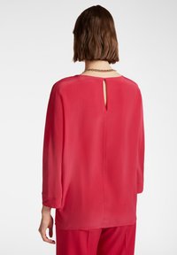 Camicia rossa trasparente con maniche a tre quarti, dettaglio a chiave sul retro e texture liscia. Abbinata a una semplice collana a catena.