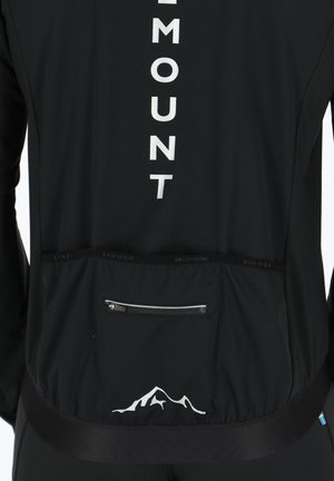 Sort sportsjakke med et sølvlogo, en lynlåslomme bagpå og en bjergsilhuet-detalje ved kanten.