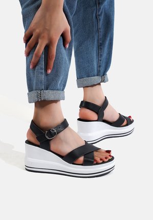Sandali wedge neri con cinturini incrociati, fibbia in argento e piattaforma bianca con strisce nere. Indossati con jeans blu risvoltati.