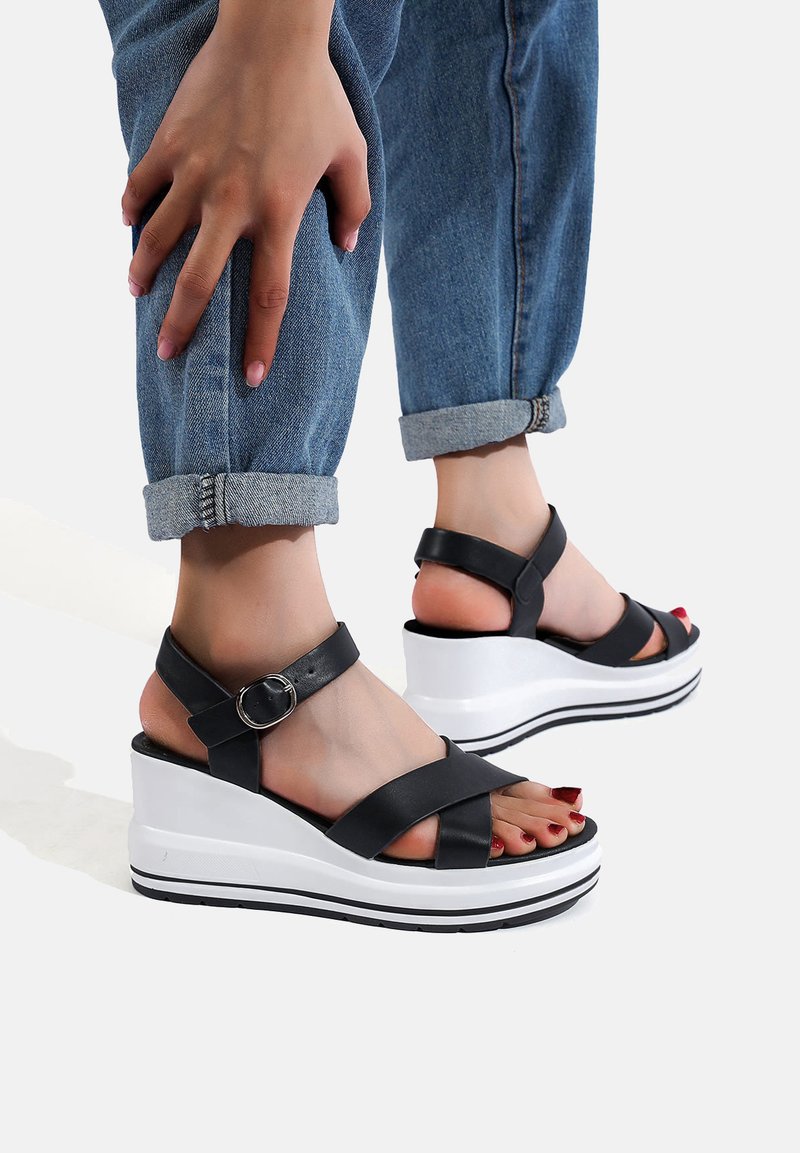 Sandali wedge neri con cinturini incrociati, fibbia in argento e piattaforma bianca con strisce nere. Indossati con jeans blu risvoltati.