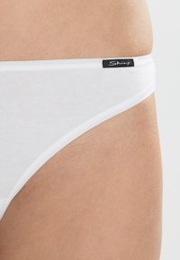Białe bawełniane bikini z gładką teksturą, z cienkim elastycznym paskiem w talii i czarną metką markową z boku.