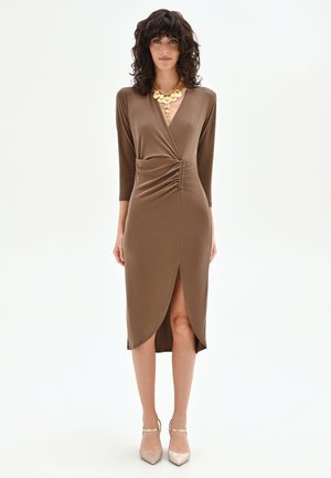 Femme aux cheveux bouclés portant une robe midi marron style portefeuille avec des détails froncés et un collier doré imposant, debout devant un fond blanc.