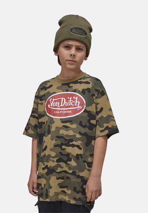 Garçon portant un bonnet vert olive et un t-shirt camouflage avec un grand logo Von Dutch California rouge et blanc sur la poitrine.