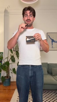 Peněženka Nike držena v ruce, černá s bílým logem, oranžový akcent, viditelné tři sloty na karty. Model má na sobě bílé tričko a modré džíny.