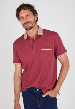 Hombre con cabello corto y rizado y barba, vistiendo una camiseta polo roja con ribete blanco, de pie con una mano en el bolsillo.