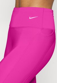 Růžové legíny s přiléhavým střihem a žebrovaným pasem, hladkého textura. Na boku je bílý logotyp Nike. Obsahují také detail boční kapsy.