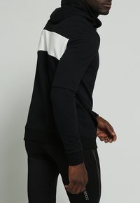 Felpa nera con cappuccio caratterizzata da un pannello orizzontale bianco contrastante sulla schiena, realizzata in materiale morbido con una finitura testurizzata e maniche aderenti.