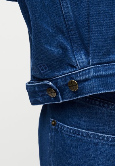 Κοντινό πλάνο στον μανσέτα μπλε τζιν μπουφάν με δύο μεταλλικά κουμπιά πάνω από τσέπη τζιν παντελονιού σε ίδιο μπλε χρώμα.