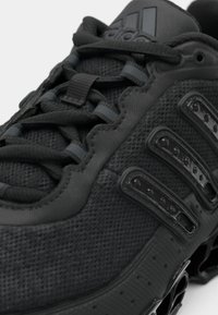 Közeli kép egy fekete Adidas edzőcipőről, amelyen texturált anyag, fekete fűzők, három fényes csík és párnázott talprészlet látható.