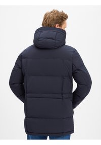 Marineblauwe pufferjas met een hoge capuchon, doorgestikt patroon en een strakke pasvorm. Heeft een rechte zoom en twee achterzakken.