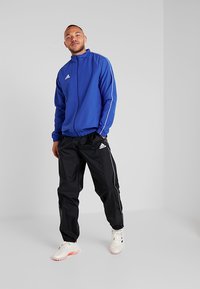 Blaue Trainingsjacke mit weißen Akzenten, Reißverschluss vorne und elastischen Bündchen, kombiniert mit schwarzen Trainingshosen mit weißen seitlichen Streifen und Kordelzug.