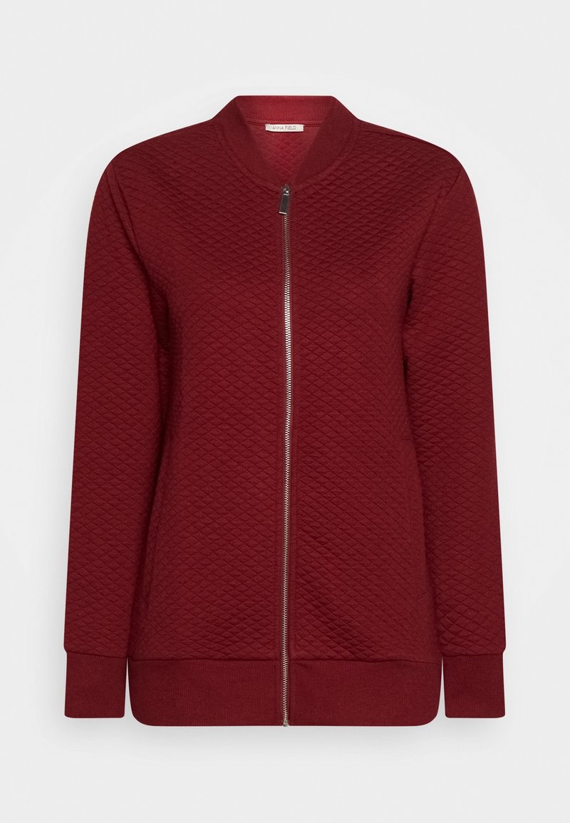 Veste bomber bordeaux avec texture matelassée, col montant, manches longues et fermeture éclair argentée sur le devant. Finition en tissu lisse.