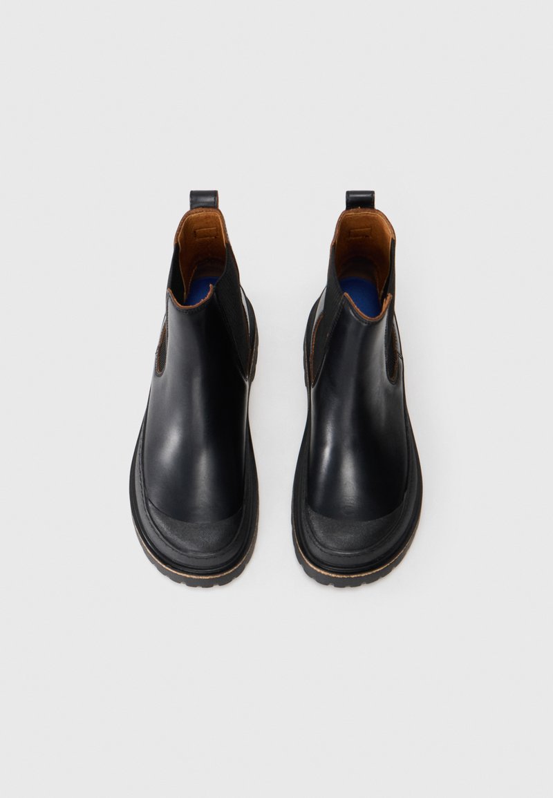 Une paire de bottes Chelsea en cuir noir avec une doublure intérieure marron et des panneaux latéraux élastiques noirs sur un fond blanc.