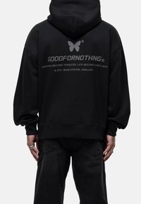 Persona con sudadera negra con logo de mariposa y el texto "GOODFORNOTHING" en la parte de atrás, combinada con pantalones negros y anillos de plata en las manos.