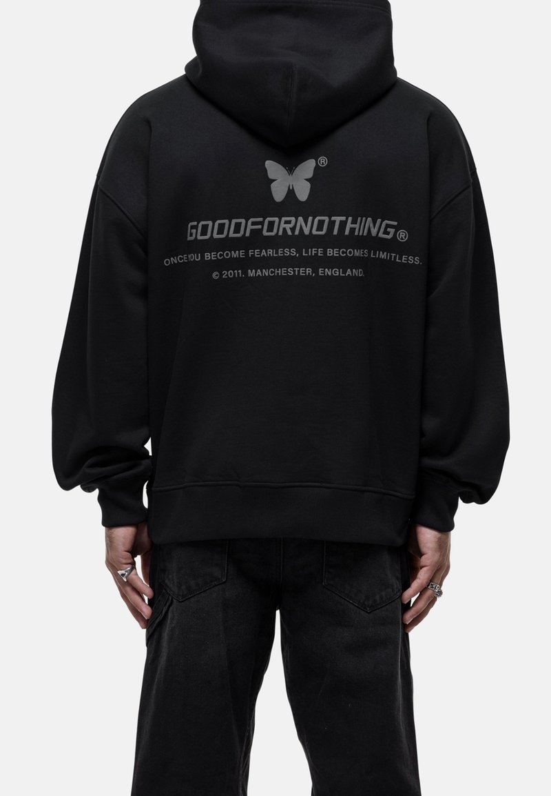 Persona con sudadera negra con logo de mariposa y el texto "GOODFORNOTHING" en la parte de atrás, combinada con pantalones negros y anillos de plata en las manos.