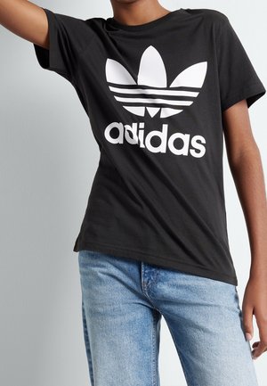 Person iført en sort Adidas t-shirt med hvidt logo og lyseblå jeans, beskåret for at vise torso og en del af armene.