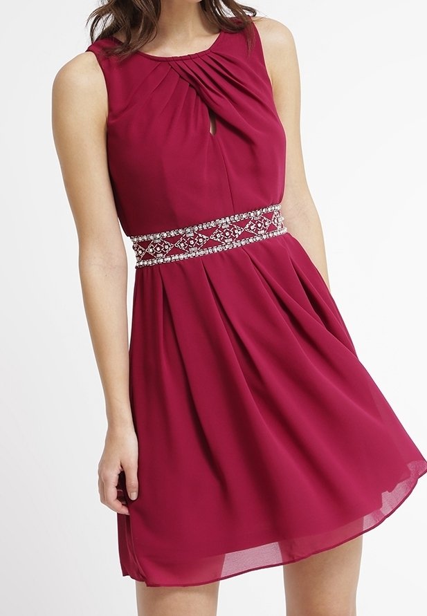 Robe rouge sans manches avec devant plissé, encolure goutte d'eau et ceinture perlée à la taille, portée par une femme debout sur fond blanc.