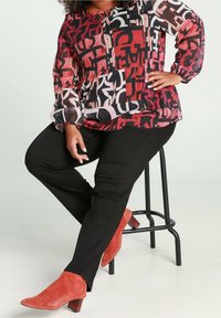 Personne assise sur un tabouret noir portant un pantalon noir, des bottes en daim rouges à talons et un chemisier à manches longues avec des motifs abstraits rouges, noirs et roses.