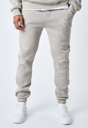 Person trägt beige Jogginghosen mit elastischen Bündchen und weißen Sneakers, steht mit den Händen in den Taschen vor einem schlichten Hintergrund.