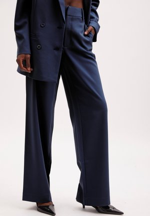 Vrouw die marineblauwe wijde broek en bijpassend dubbelregelig colbert draagt, gecombineerd met zwarte pumps met spitse neus, hand in de zak.