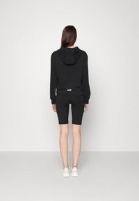 Svart hoodie med huva och logotyp på baksidan, tillsammans med svarta cykelshorts. Bärs med vita sneakers, mot en enkel bakgrund.