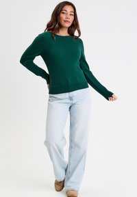 Pull en tricot vert avec un motif texturé, encolure ronde et manches longues, associé à un jean large bleu clair et des chaussures beige.