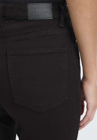 Jeans de mezclilla negra con un corte recto, que presentan un bolsillo parche y detalles de costura sutil; la etiqueta dice "PULZ JEANS."