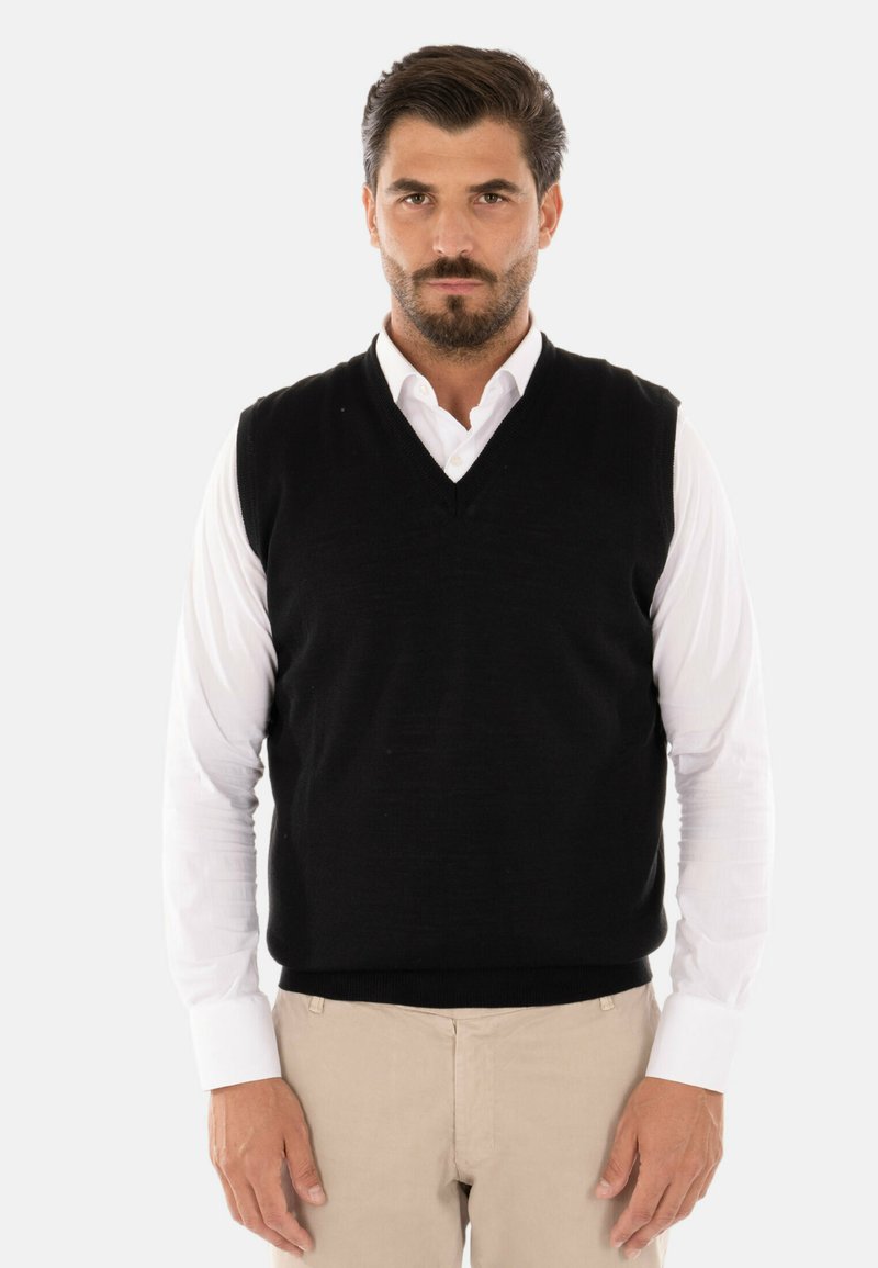 Gilet nero lavorato a maglia con scollo a V, indossato sopra una camicia bianca con bottoni, abbinato a pantaloni chiari. Texture liscia, stile aderente.