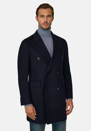 Homme portant un manteau croisé bleu marine foncé sur un pull à col roulé bleu clair, sur un fond uni.