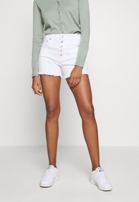 Pantalones cortos de mezclilla blanca de cintura alta con dobladillo deshilachado, que cuentan con cinco botones, combinados con una camiseta verde de manga larga y zapatillas blancas.