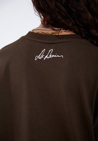 Braunes Baumwoll-T-Shirt mit einer weißen Unterschrift auf der Rückseite. Der Stoff ist glatt und im klassischen Rundhals-Design.