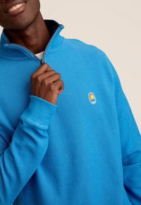 Blauwe fleece pullover met hoge kraag en half-rits sluiting. Voorzien van een badge met zon- en golvenontwerp. Geribde manchetten. Gestructureerde stof.