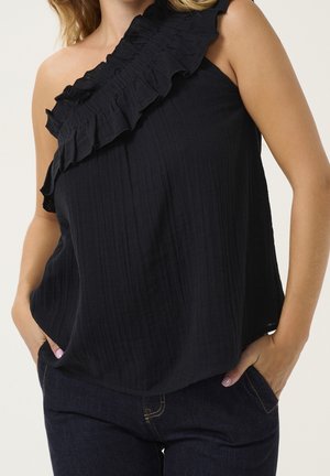 Vrouw draagt een mouwloze zwarte top met een geruffleerde single-shoulder halslijn en donkerblauwe jeans met handen in de zakken.